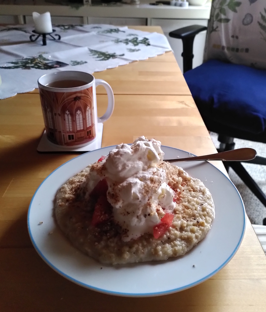 Ein Teller mit Porridge und einem großen Klecks Schlagsahne, dahinter ein Becher mit Tee.
