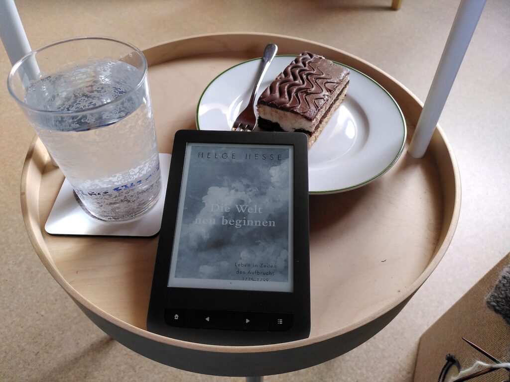 Ein Tischchen mit einem Kuchenteller, einem Glas mit Wasser und einem e-Reader. Darauf der Titel: "Die Welt neu beginnen" von Helge Hesse.