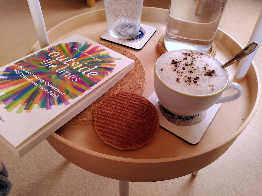 Ein kleiner Beistelltisch mit einer Tasse mit Cappuccino, einer runden Waffel, einer Flasche Wasser, einem Glas und dem Buch "Outside the lines".