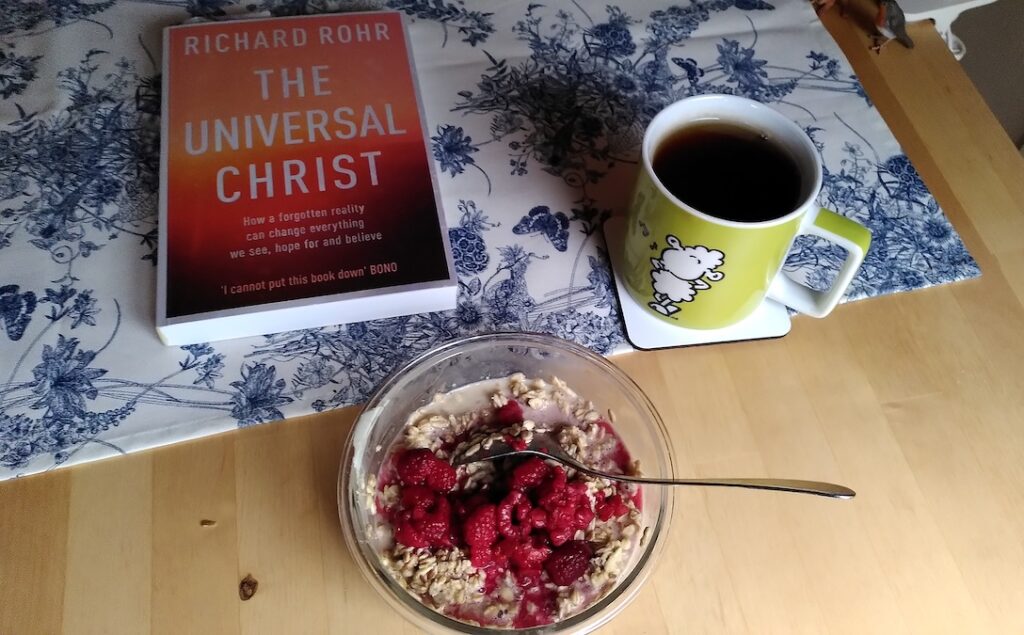 Eine Schüssel Müsli mit Himbeeren, eine Tasse schwarzer Tee und das Buch "The Universal Christ" liegen auf einem Tisch mit einem geblümten Tischläufer.