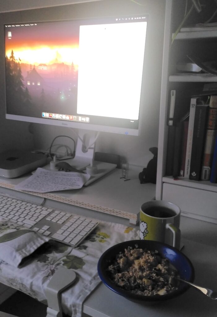 Blick auf einen Schreibtisch. Neben der Tastatur steht eine Schale mit Müsli und eine Tasse mit Tee. Auf dem Bildschirm ist auf einem märchenhaften Hintergrundbild ein weißes Textverarbeitungs-Fenster zu sehen.