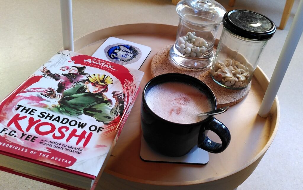 Ein Beistelltischchen mit zwei Gläsern mit Nüssen und schokolierten Beeren, einer Tasse Kakao und dem Buch "The Shadow of Kyoshi"