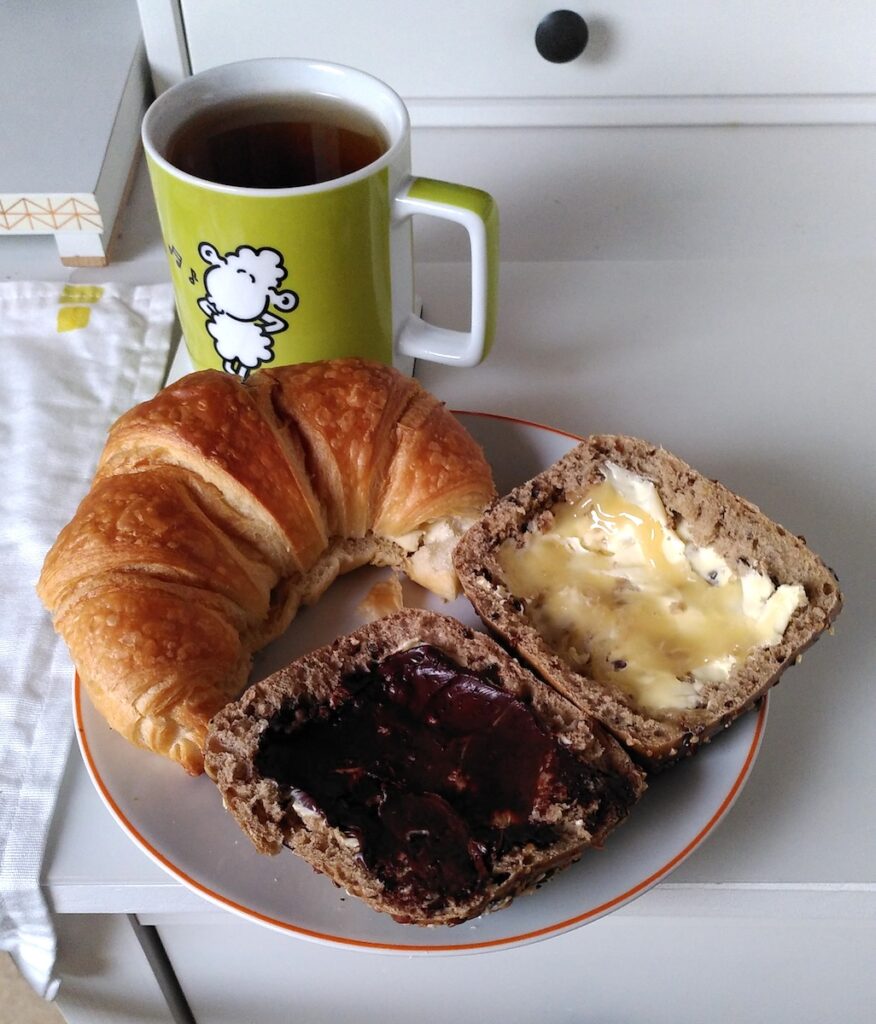 Ein Teller mit einem Croissant und zwei mit Honig und Schokocreme bestrichene Brötchenhälften auf einem Schreibtisch, dabei eine Tasse mit Schwarztee