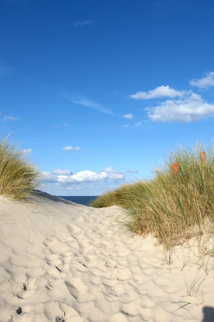 Ein Strandübergang zwischen zwei Dünen mit hohen Strandhaferpflanzen. Der helle Sand ist von Fußspuren gezeichnet, am blauen Himmel hängen ein paar Wölkchen.