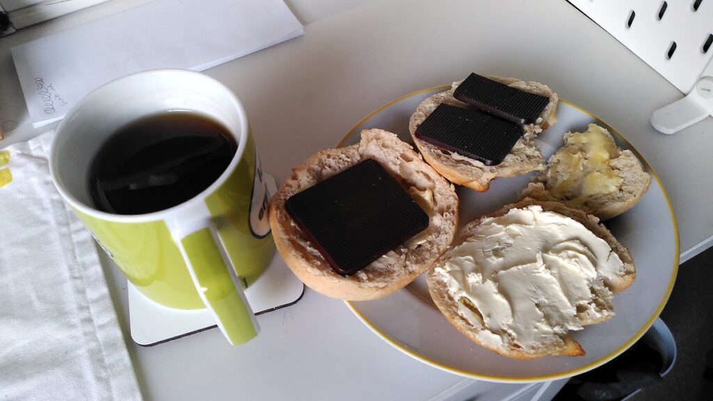 Eine Tasse mit schwarzem Tee und ein Teller mit zwei aufgeschnittenen Brötchen mit Honig, Frischkäse und Schokolade stehen auf einem Schreibtisch.