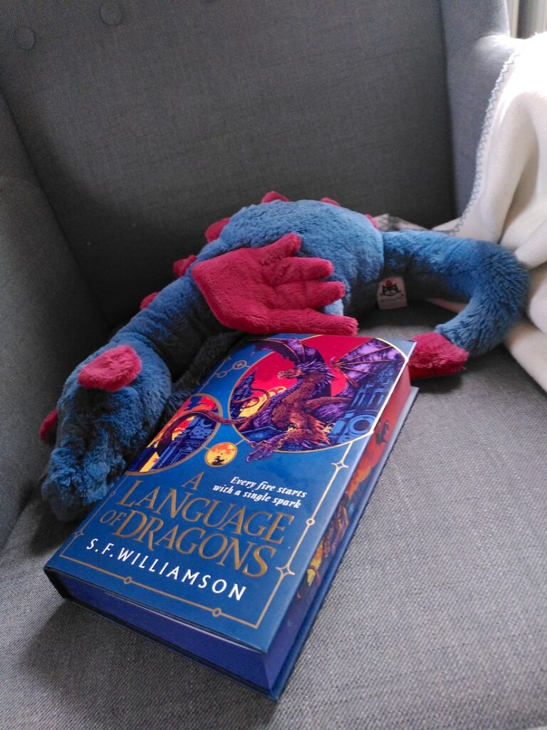 "A Language of Dragons" liegt auf einem Sessel, daneben ein Stoffdrache. Das Cover ist in dunklem Türkisblau gehalten, oben zwei runde Bilder mit einem Drachen, der vor rotem Himmel auf einem alten Gebäude sitzt. Auch auf dem Schnitt sieht man ein ähnliches Bild. Der Stoffdrache ist ebenfalls dunkeltürkis mit magentafarbenden Flügeln, Ohren, Schwanzspitze und Stacheln am Rücken.