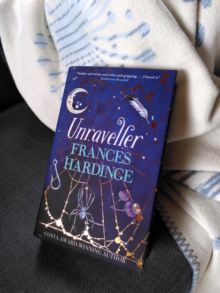 Das Buch "Unraveller" von Frances Hardinge lehnt an einer Kuscheldecke. Der Schutzumschlag ist dunkelblau mit einem goldenen Spinnennetz darauf, in dem einige Gegenstände hängen: Ein Mond, eine Feder, ein Angelhaken und Mohnkapseln, außerdem sitzt eine Spinne darin.